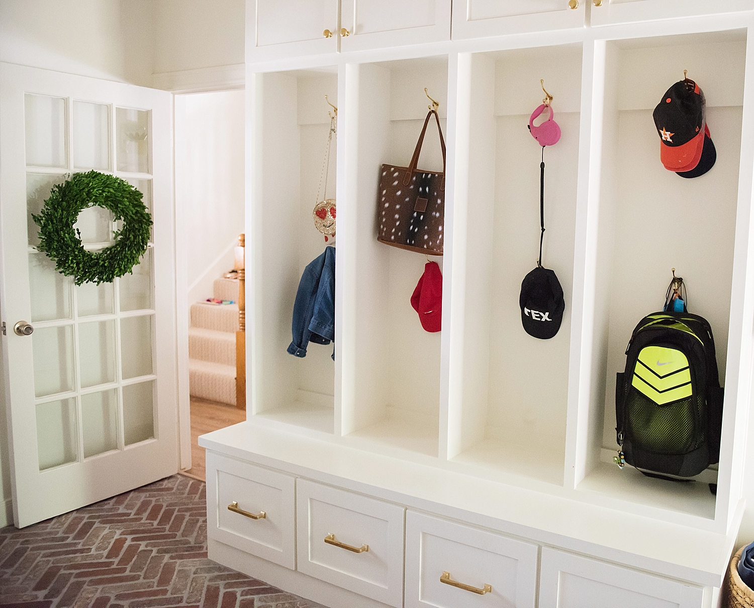 Navy Blue Paint by popular Houston life and style blog, Fancy Ashley: image of a mudroom with white cabinets, brick flooring, and gold hooks with backpacks, bags, and hats hanging from them. 