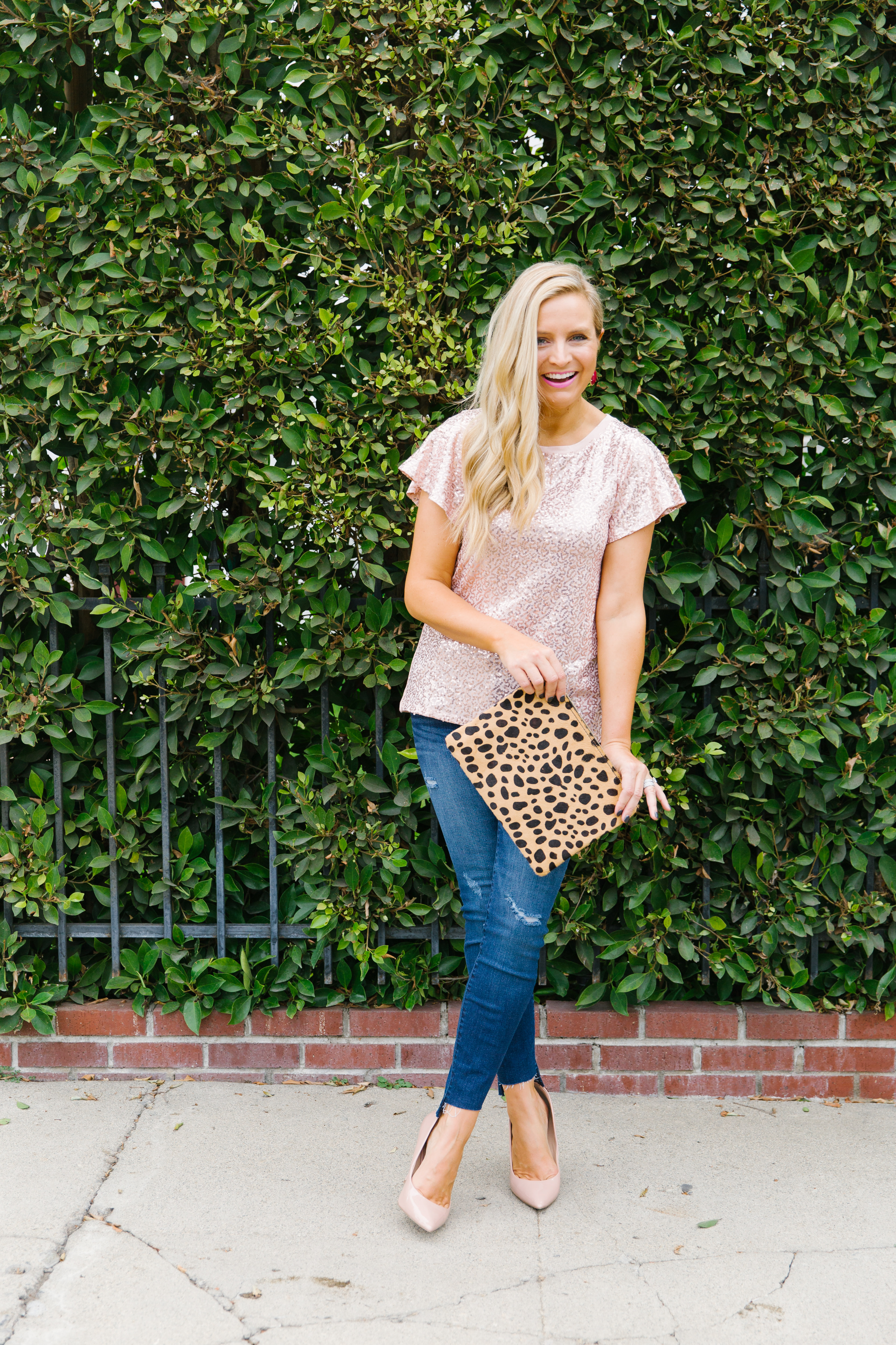 Gibson x Glam Collection Outfits featured by top Houston fashion blog, Fancy Ashley: a blonde woman walking wearing sequin flutter sleeves top, jeans, leopard clutch and pink heels
