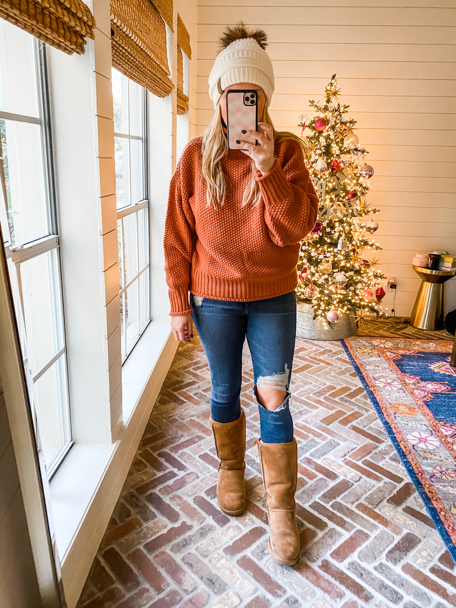 December Outfits by popular Houston fashion blog, Fancy Ashley: image of a woman standing in front of a Christmas tree decorated with pink, white and silver ornaments and wearing a orange cable knit sweater, distressed denim, cream fur pom beanie, and brown suede Ugg boots.