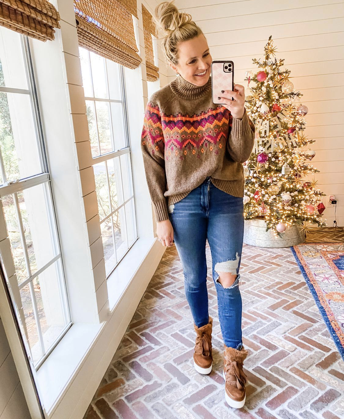 December Outfits by popular Houston fashion blog, Fancy Ashley: image of a woman standing in front of a Christmas tree decorated with pink, white and silver ornaments and wearing a brown, pink and orange nordic print turtleneck sweater, distressed denim, and brown fur lined hightop sneakers. 
