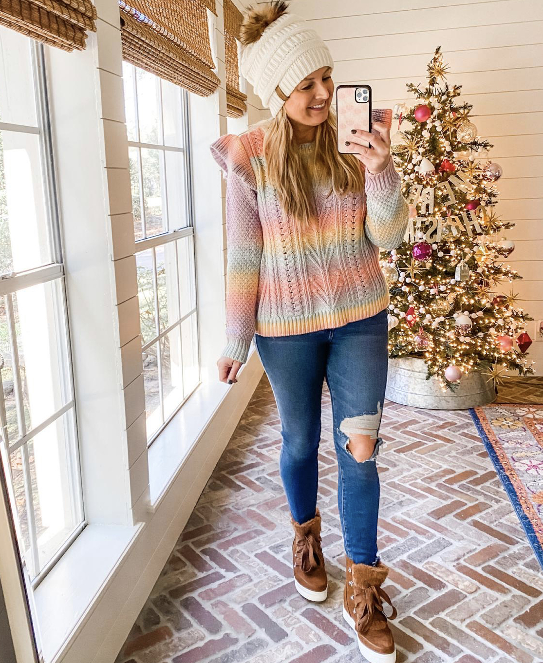 December Outfits by popular Houston fashion blog, Fancy Ashley: image of a woman standing in front of a Christmas tree decorated with pink, white and silver ornaments and wearing a multi color stripe knit sweater, cream fur pom beanie, distress denim, and brown fur lined hight tops sneakers. 