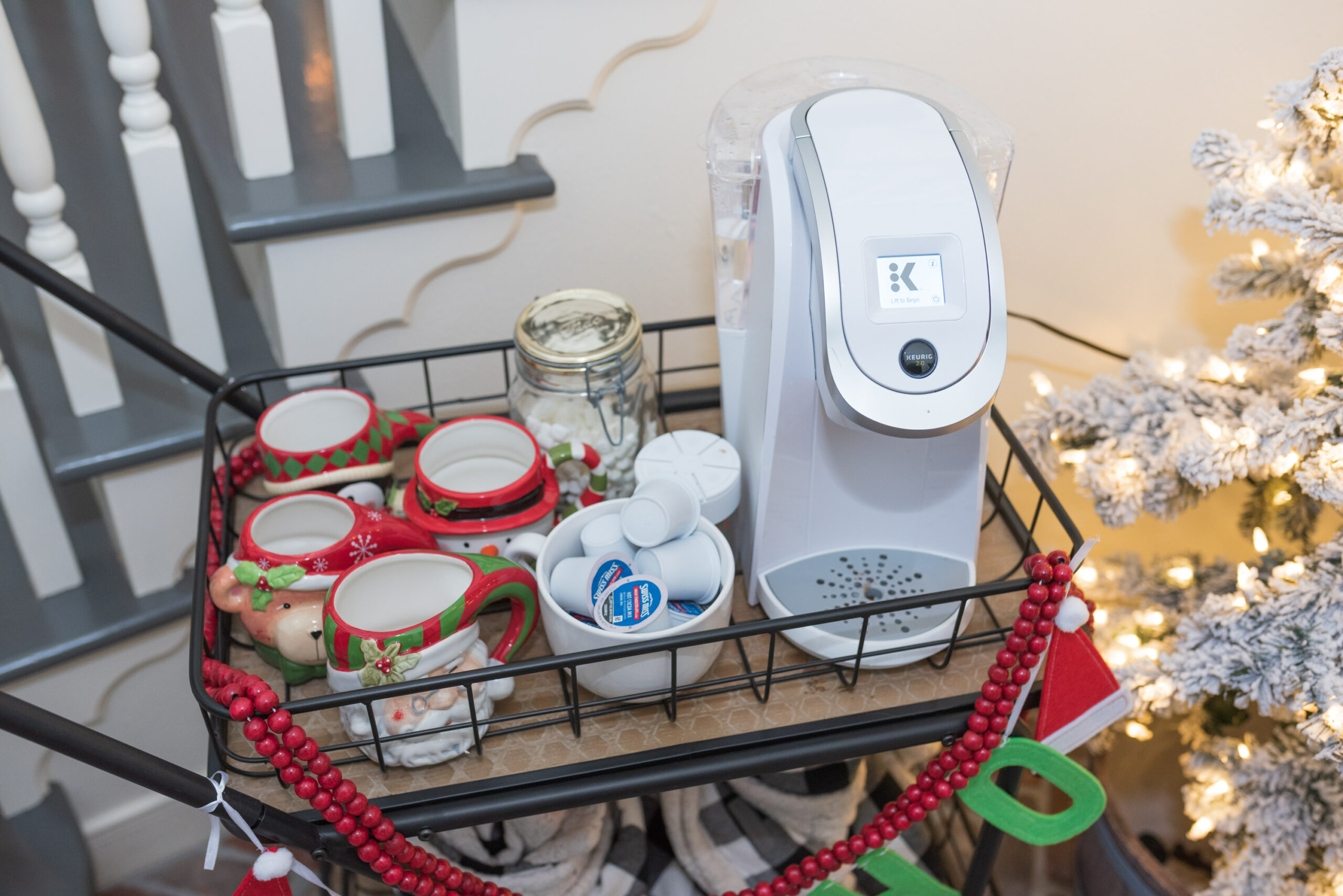 Hot Cocoa Bar by popular Houston lifestyle blog, Fancy Ashley: image of a black metal bar cart containing a Keurig, Christmas mugs, Christmas garland, mini marshmallows, and coco pods. 