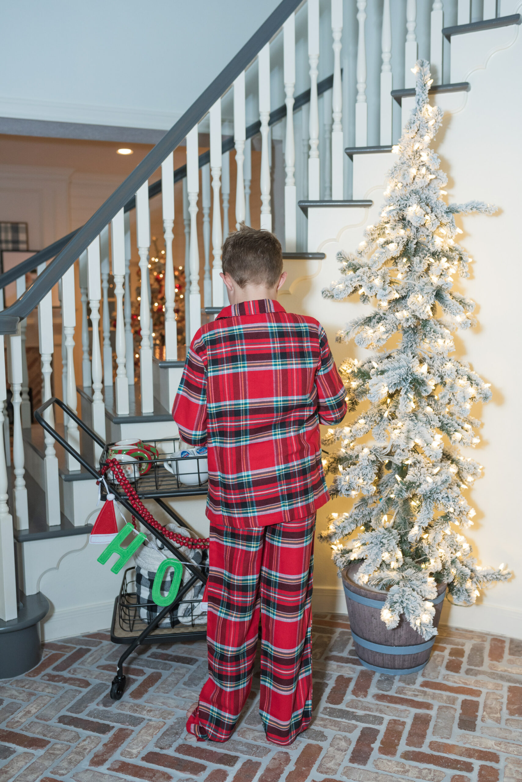 Hot Cocoa Bar by popular Houston lifestyle blog, Fancy Ashley: image of a young boy wearing plaid pajamas and standing next to a black metal bar cart containing a Keurig, Christmas mugs, Christmas garland, mini marshmallows, and coco pods. 