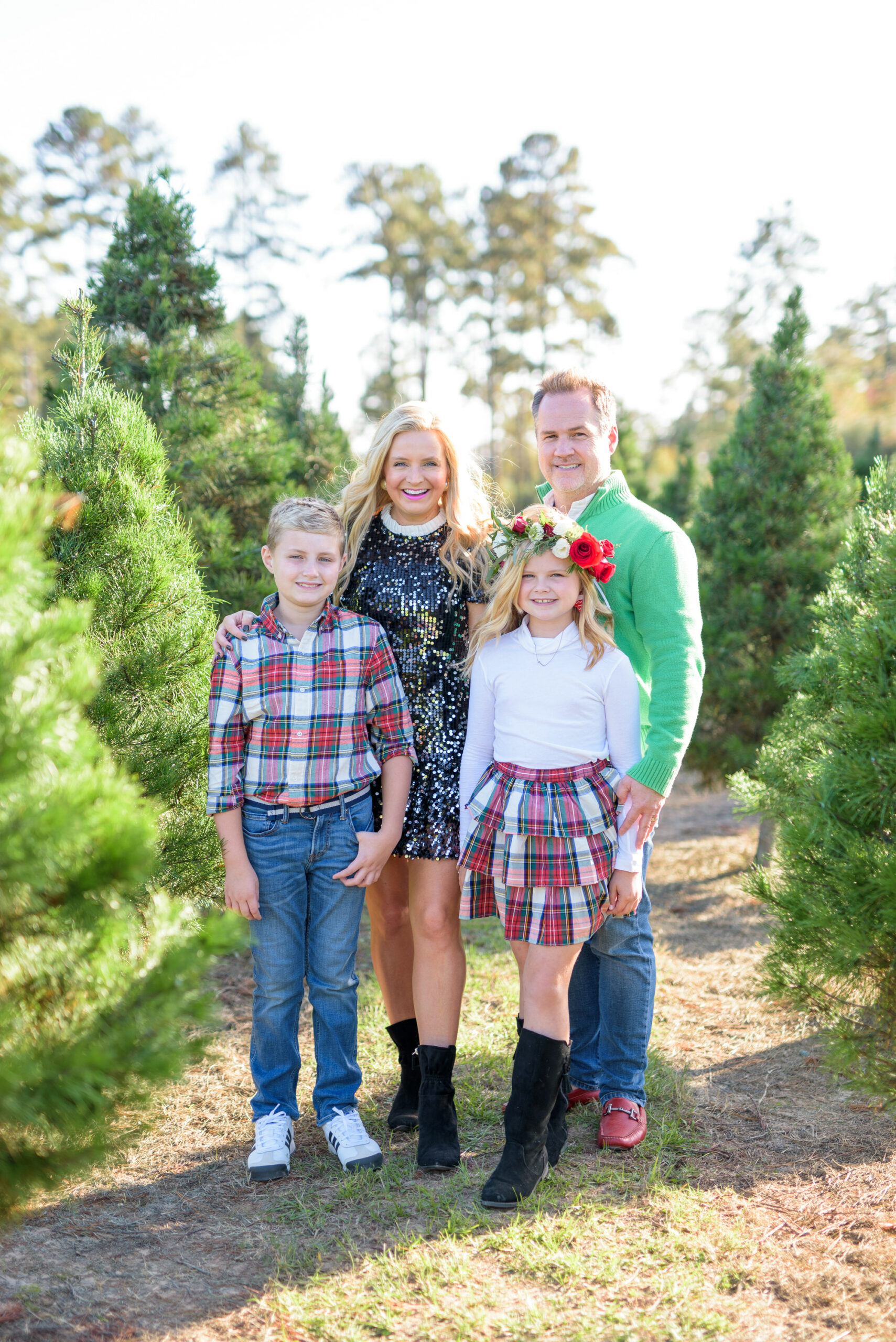 Christmas Tree Farm Photos by popular Houston lifestyle blog, Fancy Ashley: image of a family standing in a row of pine trees and wearing a black sequin dress, black suede ankle boots, plaid tier ruffle skirt, flower crown, plaid button up shirt, red loafers, and green pullover sweater. 