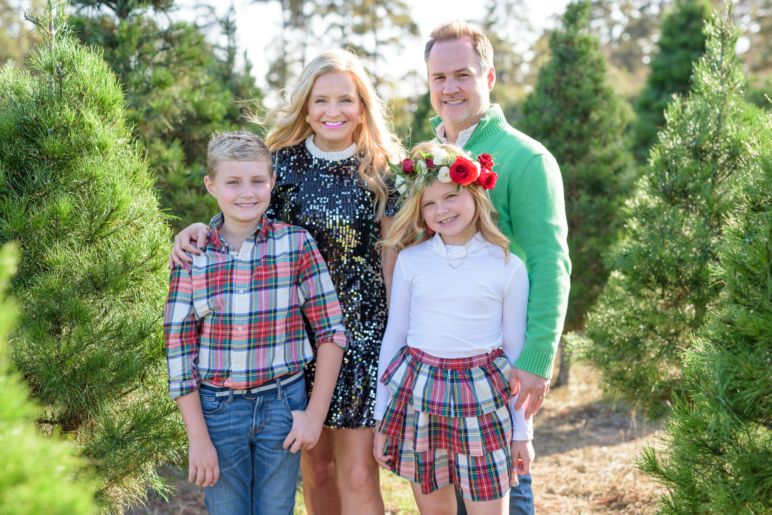 Christmas Tree Farm Photos by popular Houston lifestyle blog, Fancy Ashley: image of a family standing in a row of pine trees and wearing a black sequin dress, black suede ankle boots, plaid tier ruffle skirt, flower crown, plaid button up shirt, red loafers, and green pullover sweater. 