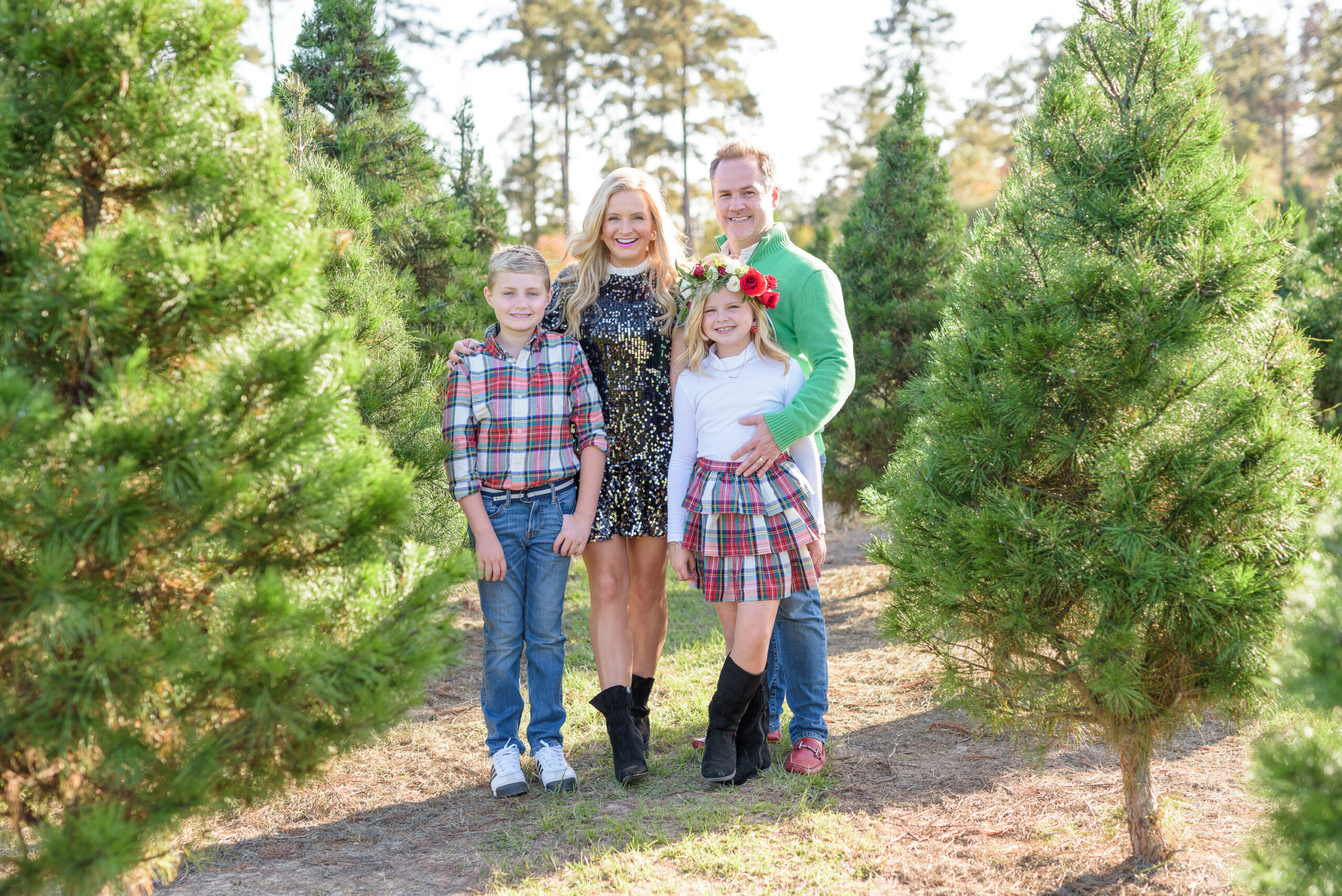 Christmas Tree Farm Photos by popular Houston lifestyle blog, Fancy Ashley: image of a family standing in a row of pine trees and wearing a black sequin dress, black suede ankle boots, plaid tier ruffle skirt, flower crown, plaid button up shirt, red loafers, and green pullover sweater. 