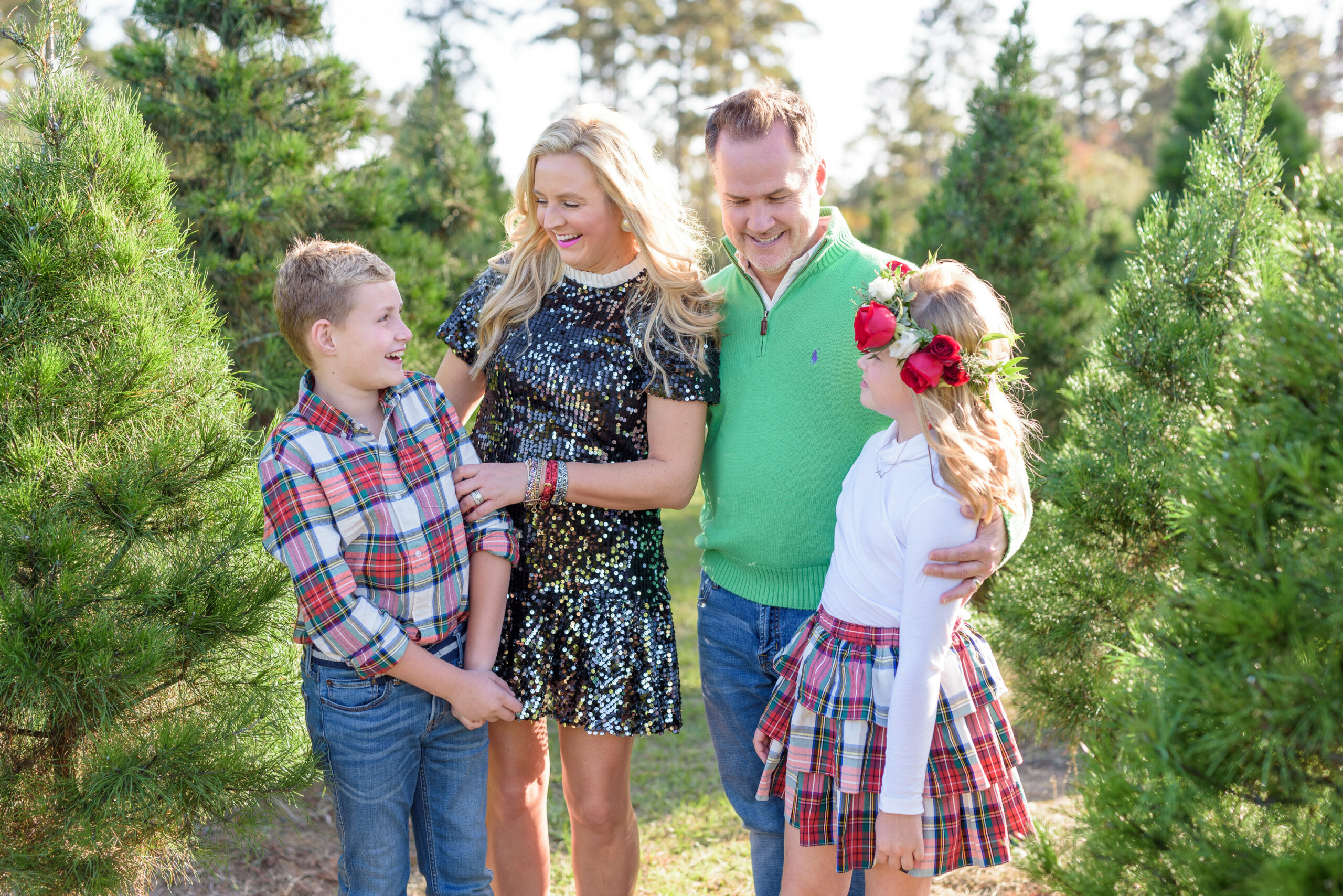 Christmas Tree Farm Photos by popular Houston lifestyle blog, Fancy Ashley: image of a family standing in a row of pine trees and wearing a black sequin dress, black suede ankle boots, plaid tier ruffle skirt, flower crown, plaid button up shirt, red loafers, and green pullover sweater. 