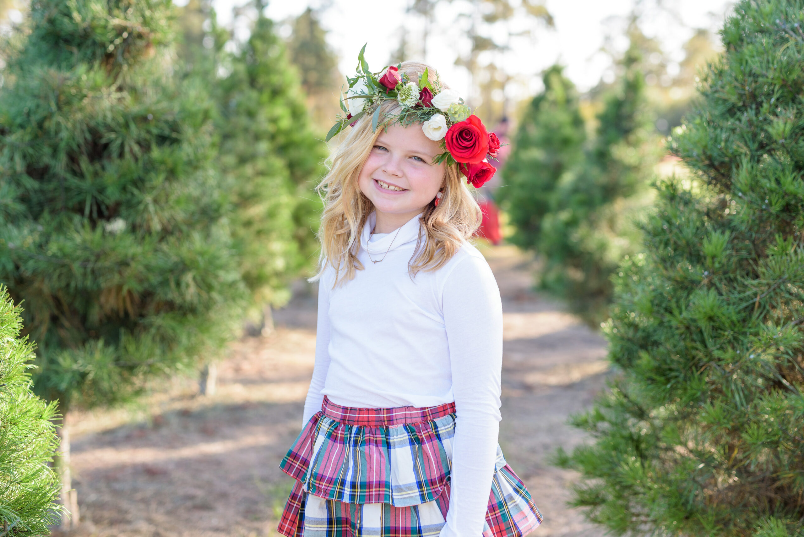 Christmas Tree Farm Photos by popular Houston lifestyle blog, Fancy Ashley: image of a girl standing in a row of pine trees and wearing a white long sleeve turtleneck, plaid tier ruffle skirt, flower crown