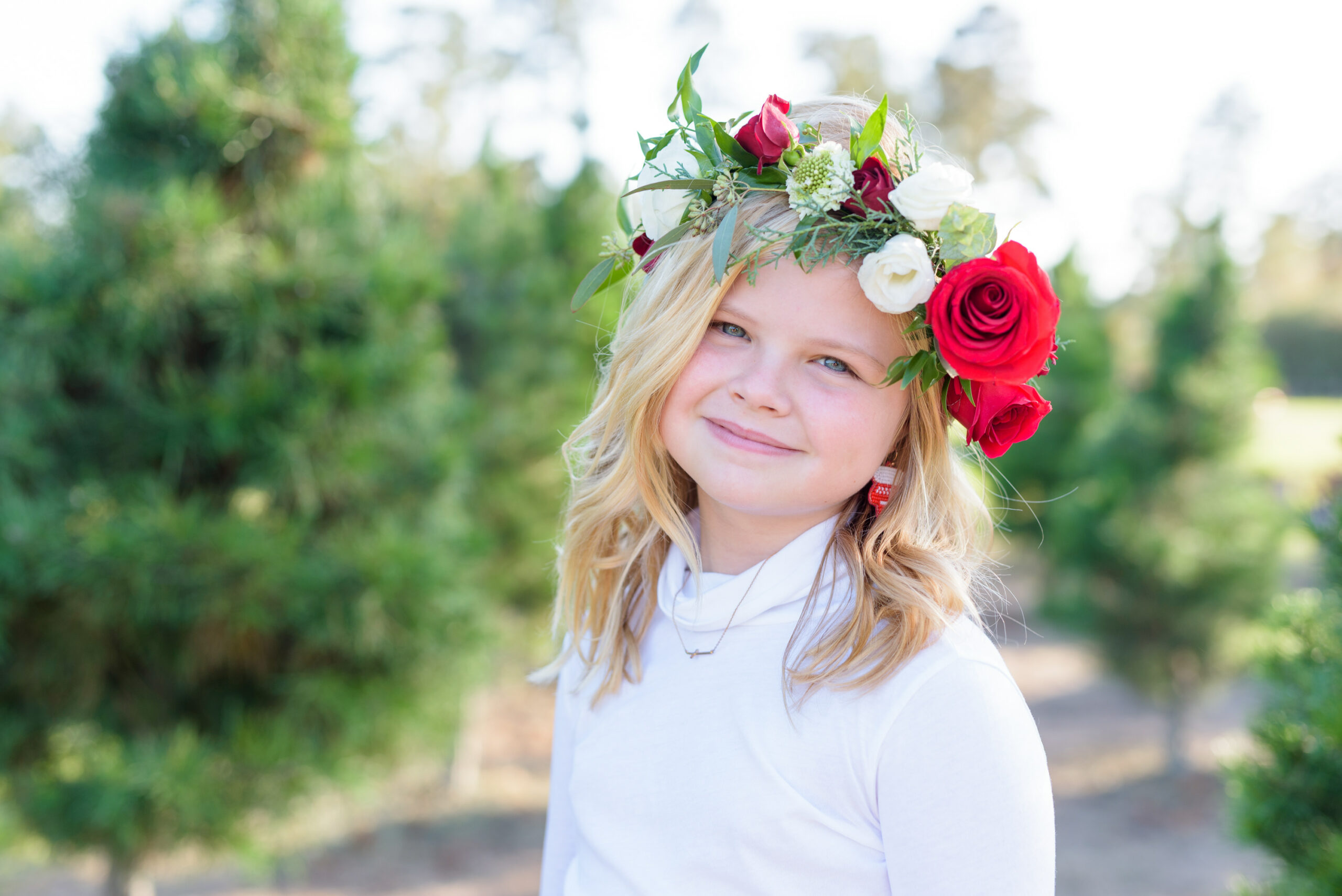 Christmas Tree Farm Photos by popular Houston lifestyle blog, Fancy Ashley: image of a girl standing in a row of pine trees and wearing a white long sleeve turtleneck, plaid tier ruffle skirt, flower crown