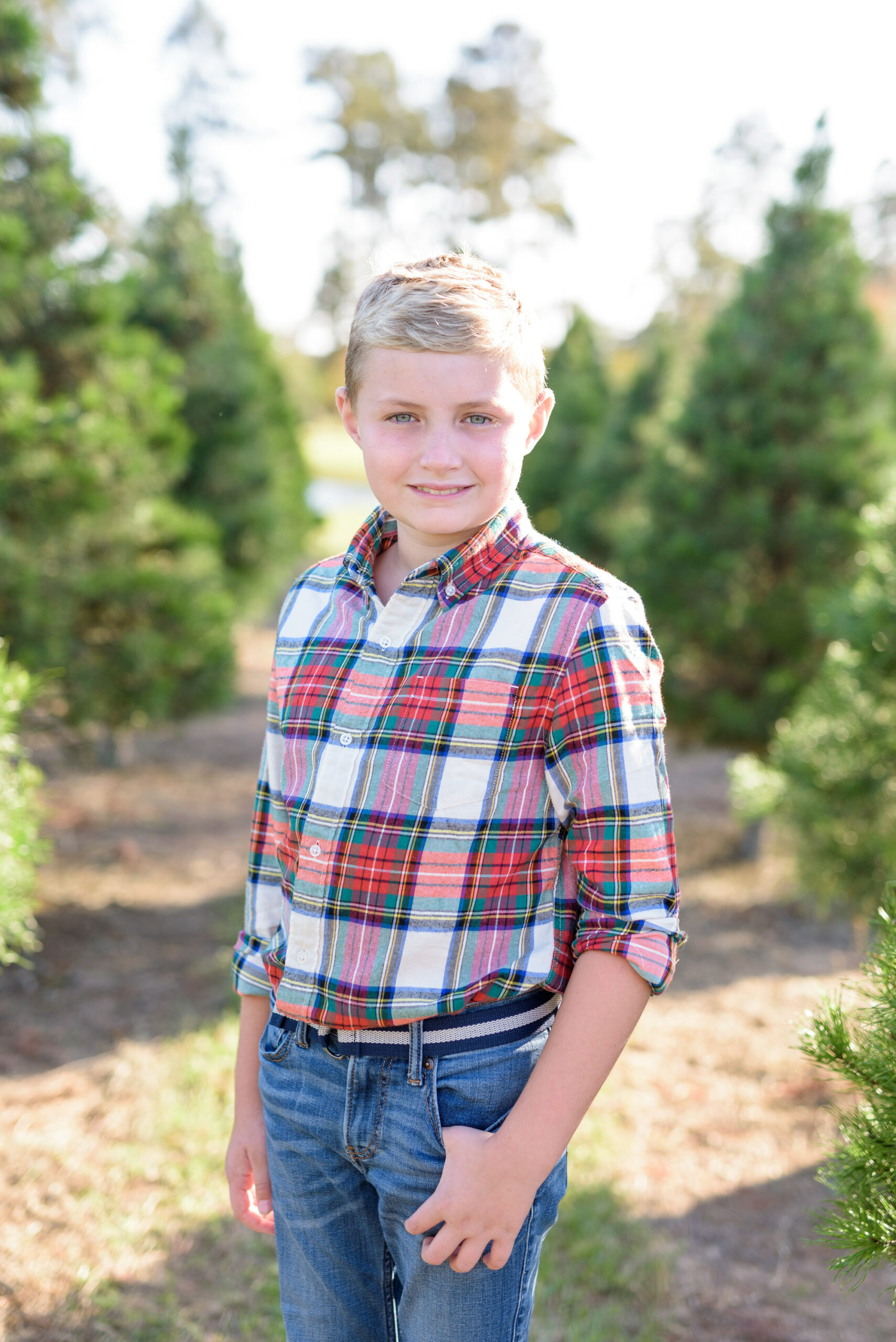 Christmas Tree Farm Photos by popular Houston lifestyle blog, Fancy Ashley: image of a boy standing in a row of pine trees and wearing a plaid button up shirt and jeans. 