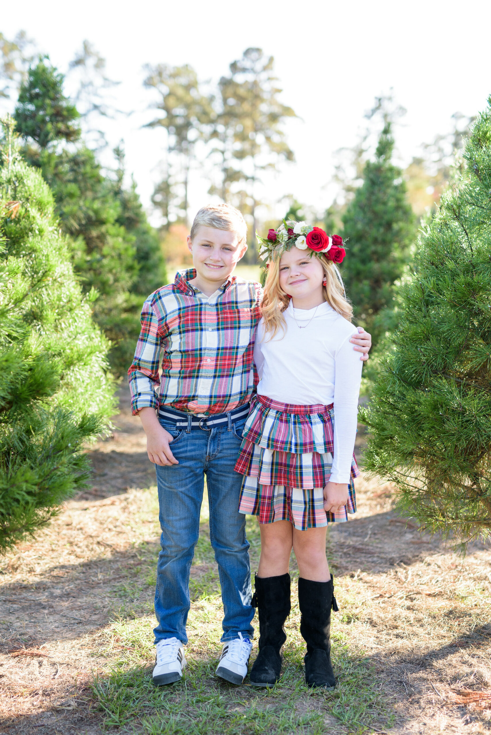 Christmas Tree Farm Photos by popular Houston lifestyle blog, Fancy Ashley: image of a young boy and girl standing together in a row of pine trees and wearing a white long sleeve turtleneck shirt, plaid tier ruffle skirt, flower crown, plaid button up shirt, and jeans. 