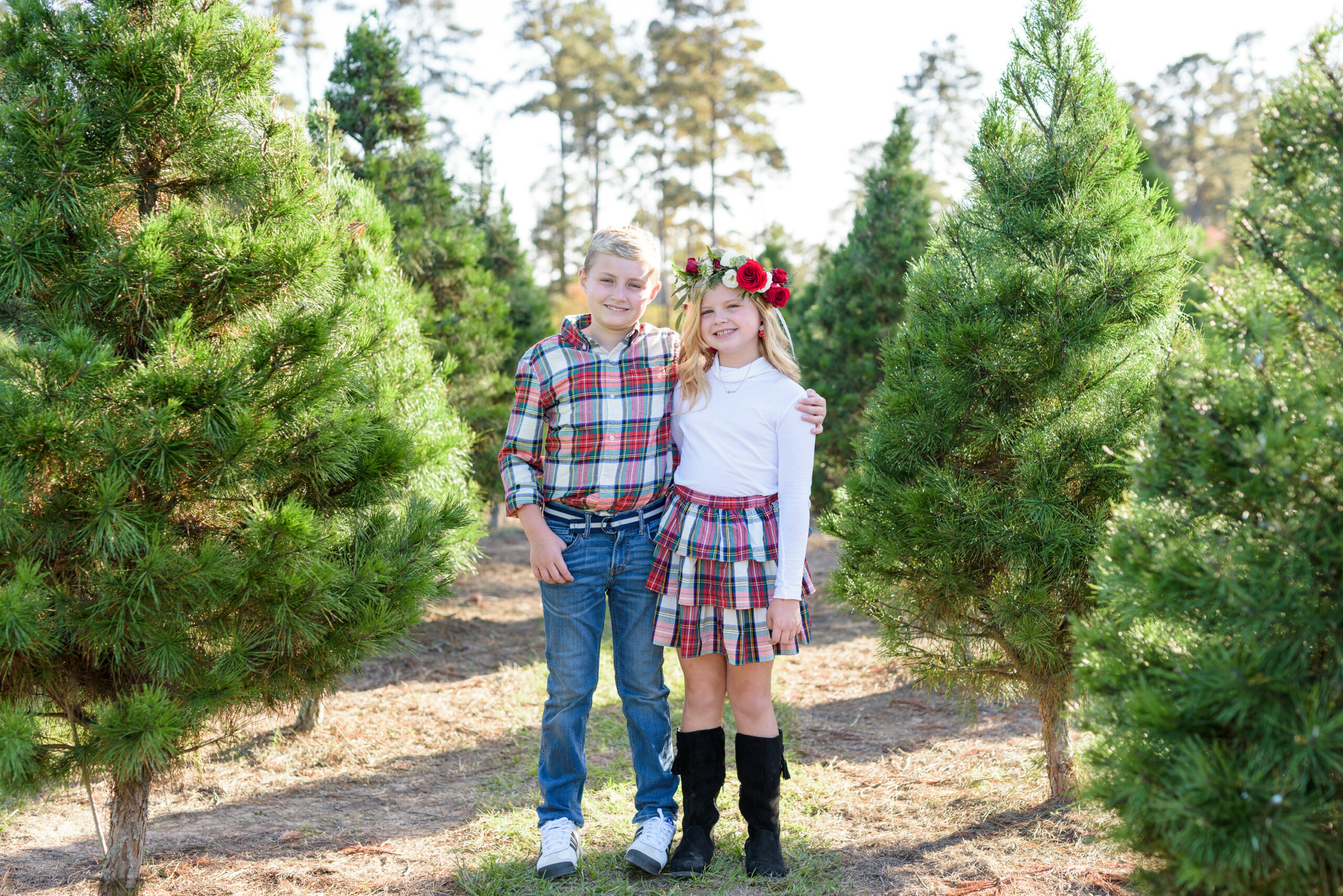 Christmas Tree Farm Photos by popular Houston lifestyle blog, Fancy Ashley: image of a young boy and girl standing together in a row of pine trees and wearing a white long sleeve turtleneck shirt, plaid tier ruffle skirt, flower crown, plaid button up shirt, and jeans. 