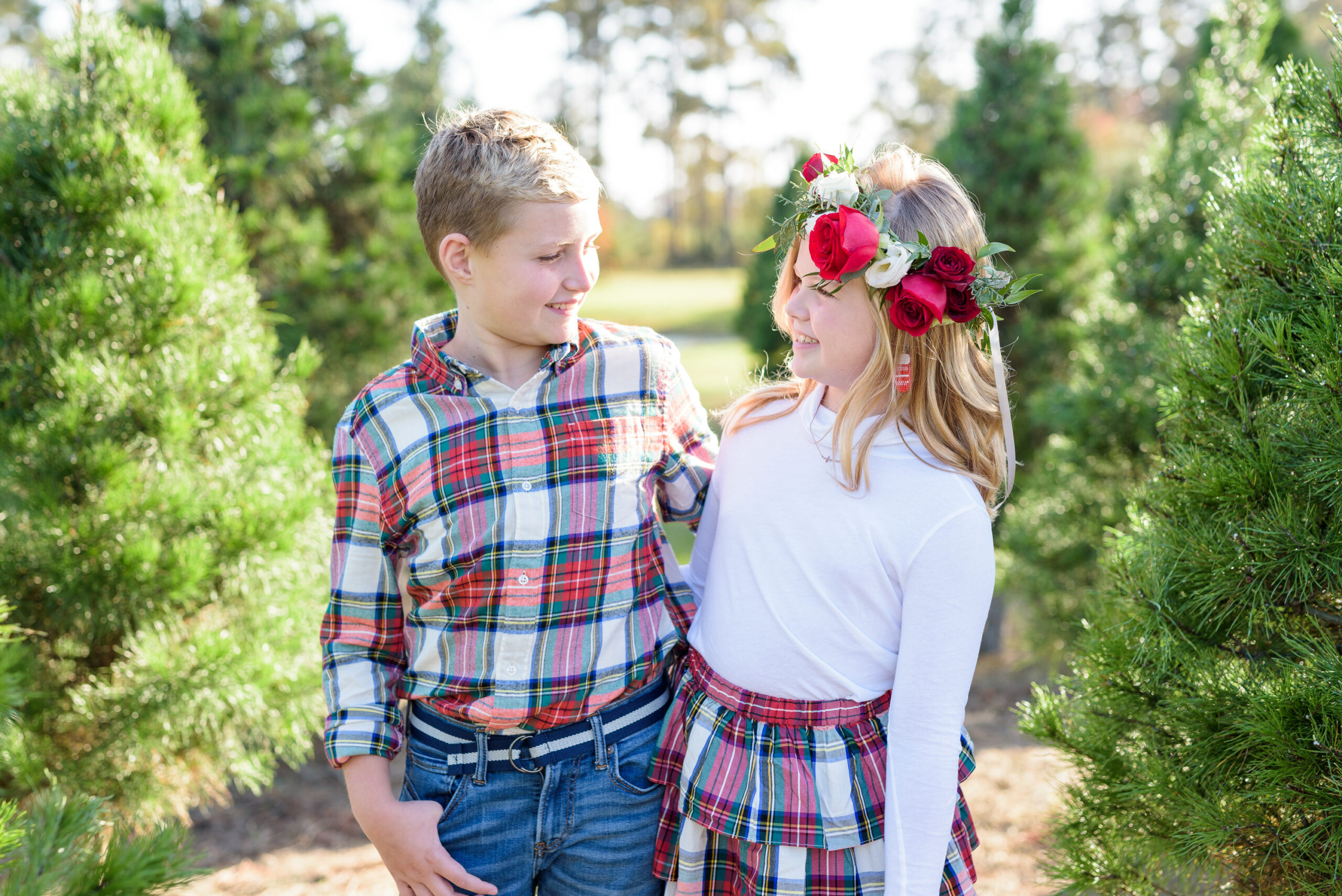 Christmas Tree Farm Photos by popular Houston lifestyle blog, Fancy Ashley: image of a young boy and girl standing together in a row of pine trees and wearing a white long sleeve turtleneck shirt, plaid tier ruffle skirt, flower crown, plaid button up shirt, and jeans. 