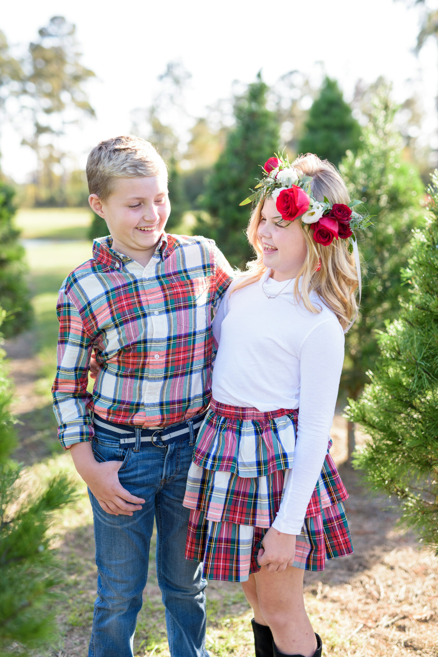 Christmas Tree Farm Photos by popular Houston lifestyle blog, Fancy Ashley: image of a young boy and girl standing together in a row of pine trees and wearing a white long sleeve turtleneck shirt, plaid tier ruffle skirt, flower crown, plaid button up shirt, and jeans. 