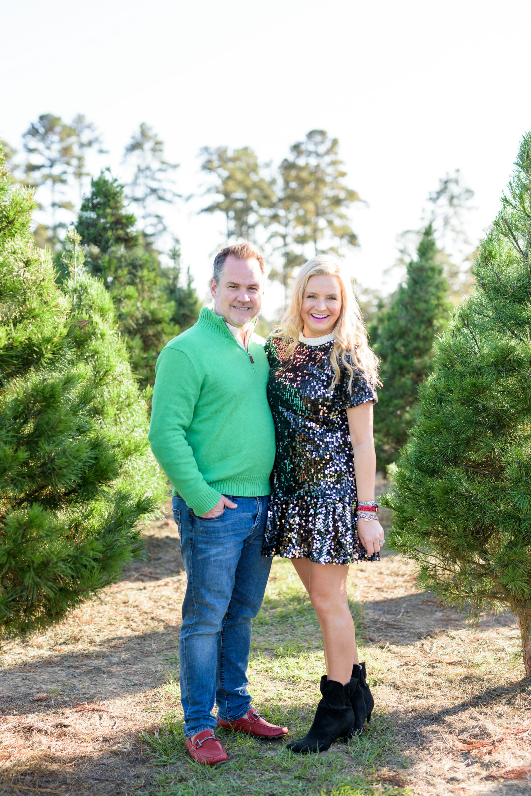 Christmas Tree Farm Photos by popular Houston lifestyle blog, Fancy Ashley: image of a husband and wife standing together in a row of pine trees and wearing a black sequin dress, black suede ankle boots, jeans, red loafers, and green pull over sweater. 