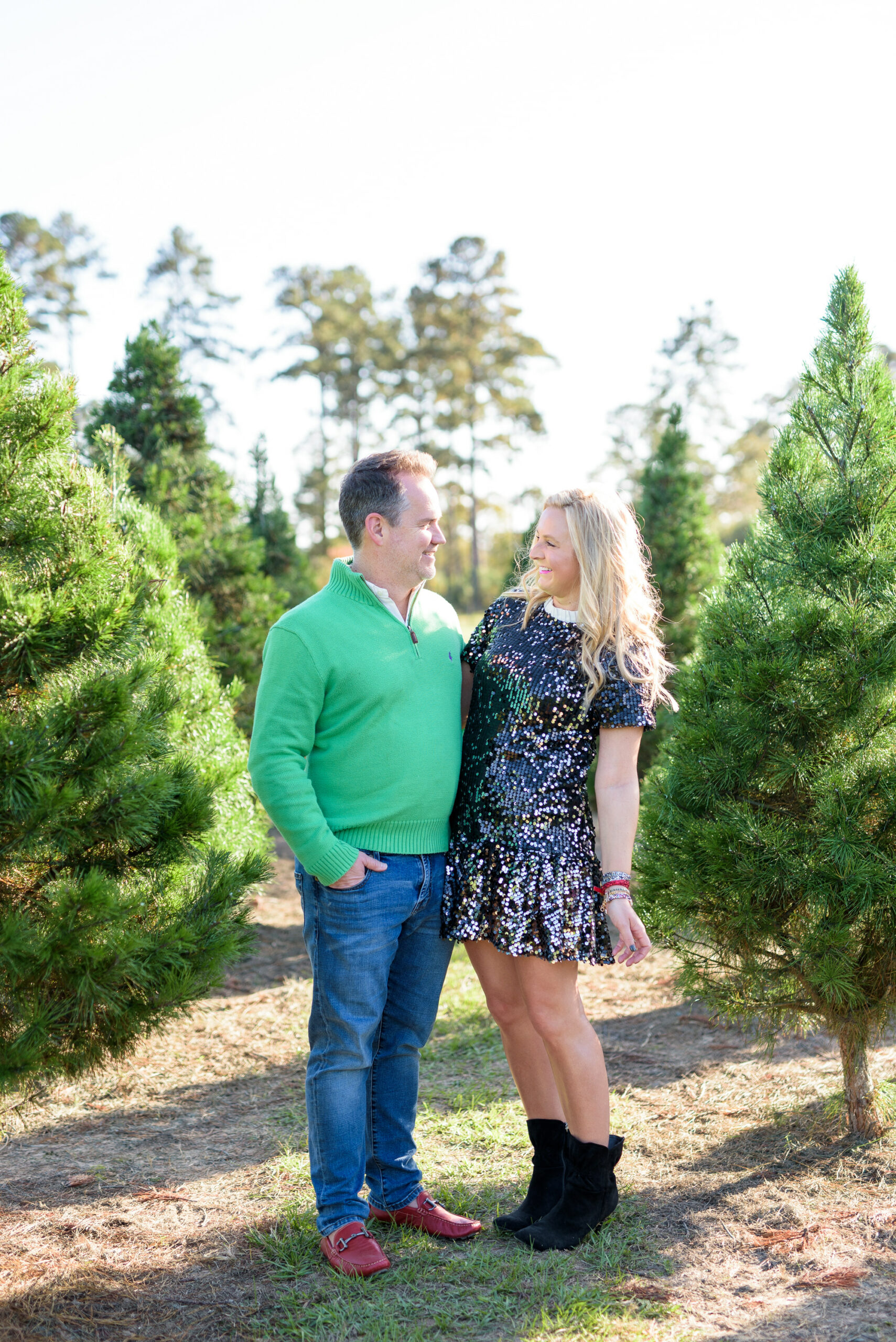 Christmas Tree Farm Photos by popular Houston lifestyle blog, Fancy Ashley: image of a husband and wife standing together in a row of pine trees and wearing a black sequin dress, black suede ankle boots, jeans, red loafers, and green pull over sweater. 