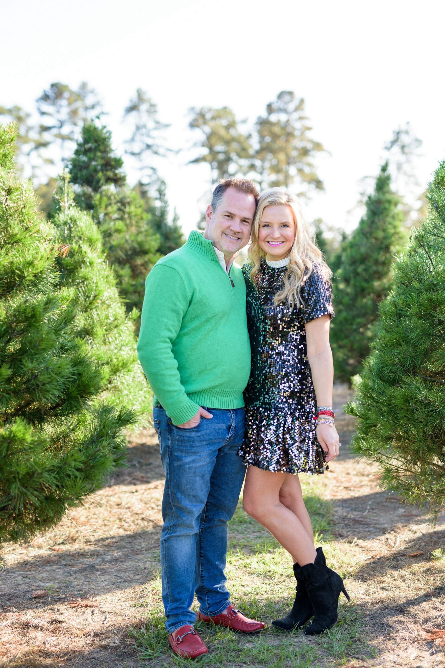 Christmas Tree Farm Photos by popular Houston lifestyle blog, Fancy Ashley: image of a husband and wife standing together in a row of pine trees and wearing a black sequin dress, black suede ankle boots, jeans, red loafers, and green pull over sweater. 