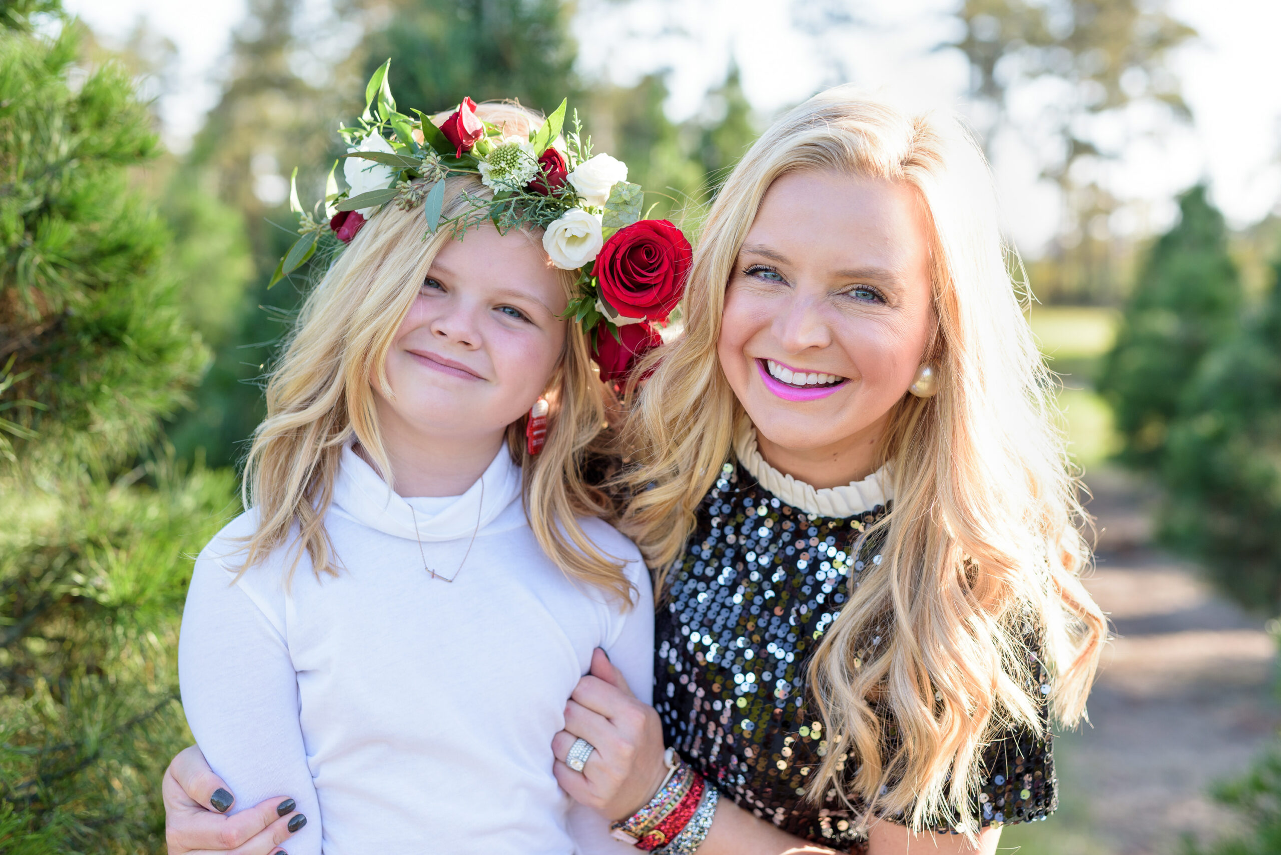 Christmas Tree Farm Photos by popular Houston lifestyle blog, Fancy Ashley: image of a mom and daughter standing together in a row of pine trees and wearing a black sequin dress, black suede ankle boots, plaid tier skirt, white long sleeve turtleneck shirt, and floral crown. 