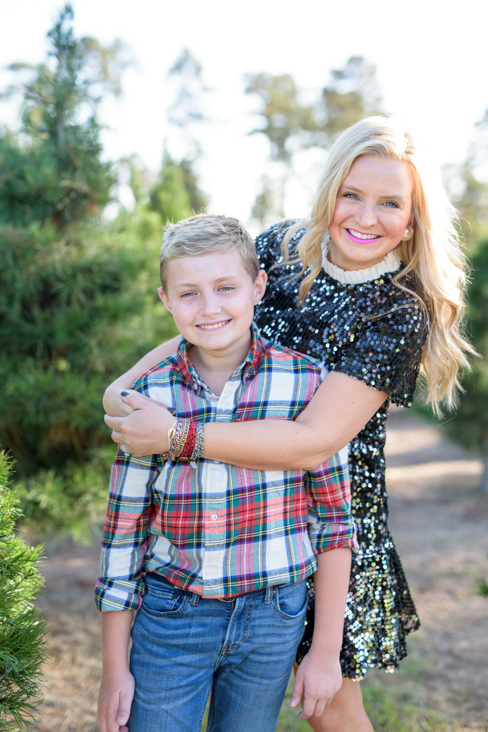 Christmas Tree Farm Photos by popular Houston lifestyle blog, Fancy Ashley: image of a mom and son standing together in a row of pine trees and wearing a black sequin dress, black suede ankle boots, plaid button up shirt, and jeans. 