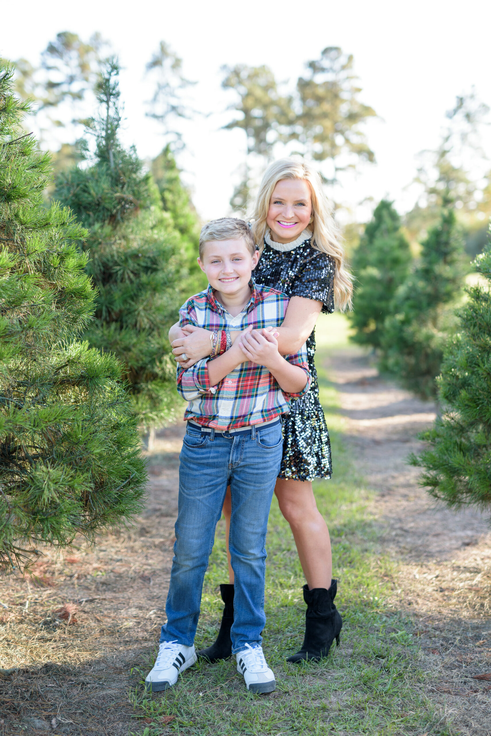 Christmas Tree Farm Photos by popular Houston lifestyle blog, Fancy Ashley: image of a mom and son standing together in a row of pine trees and wearing a black sequin dress, black suede ankle boots, plaid button up shirt, and jeans. 