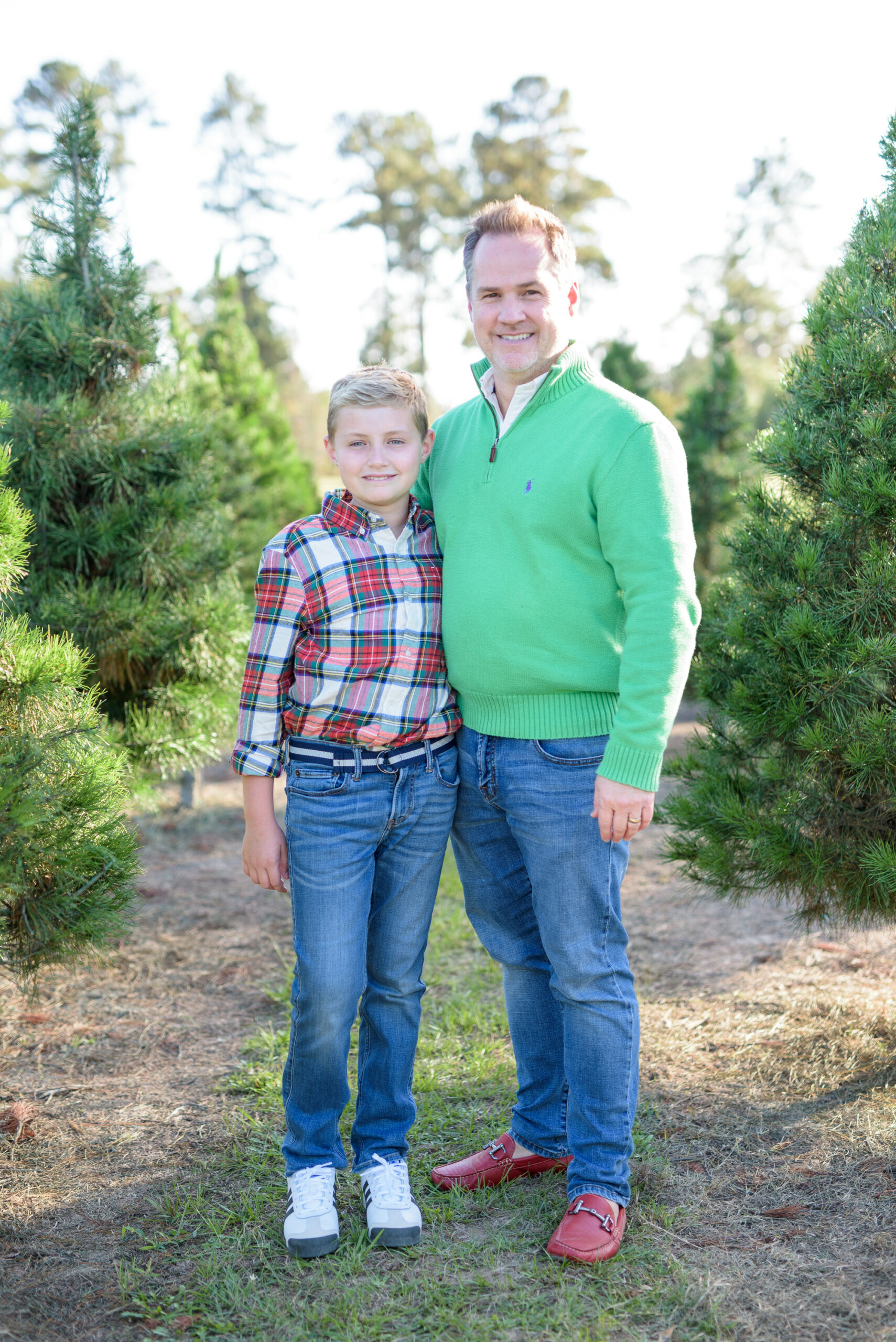 Christmas Tree Farm Photos by popular Houston lifestyle blog, Fancy Ashley: image of a dad and his daughter standing together in a row of pine trees and wearing a green pullover sweater, red loafers, plaid button up shirt, and jeans. 