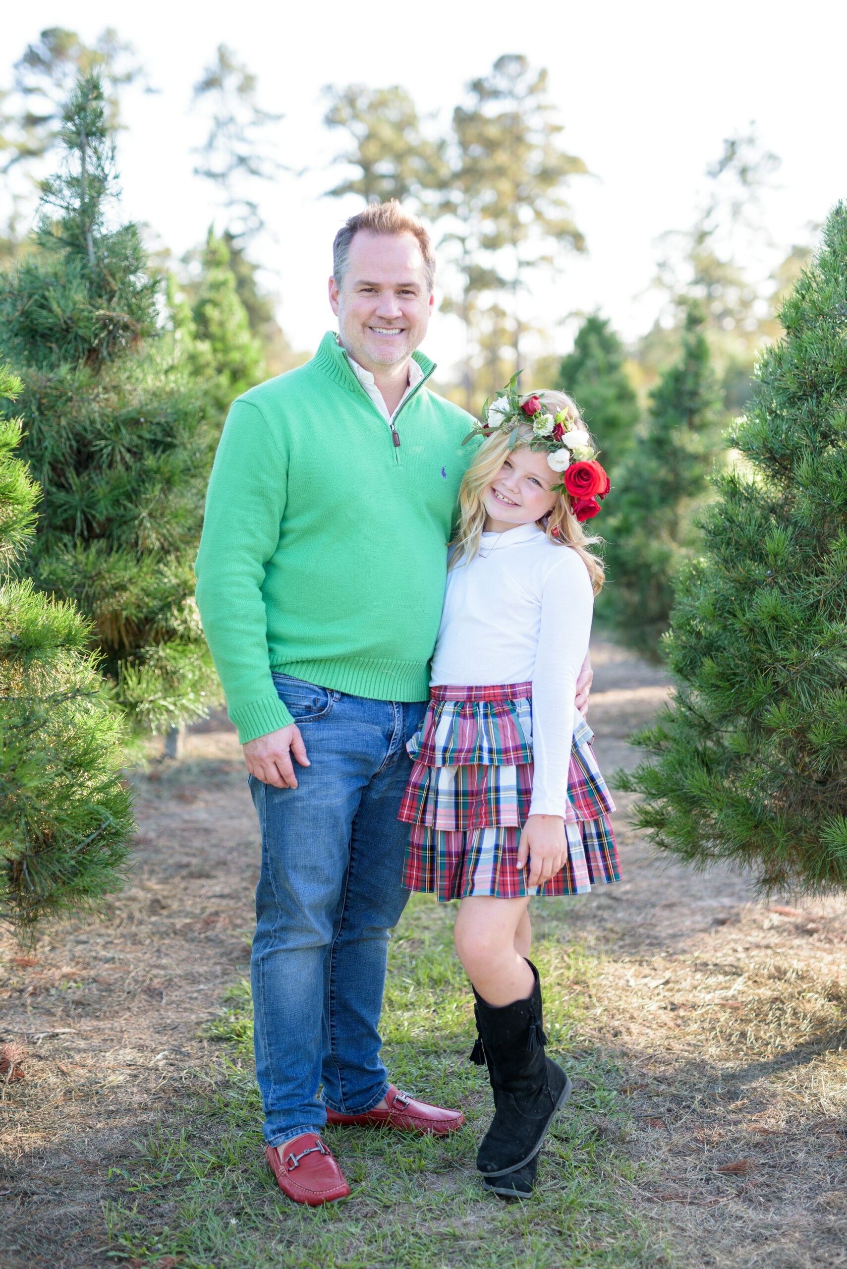 Christmas Tree Farm Photos by popular Houston lifestyle blog, Fancy Ashley: image of a dad and his daughter standing together in a row of pine trees and wearing a green pullover sweater, white long sleeve turtleneck shirt, plaid ruffle tier skirt, floral crown and black suede boots. 