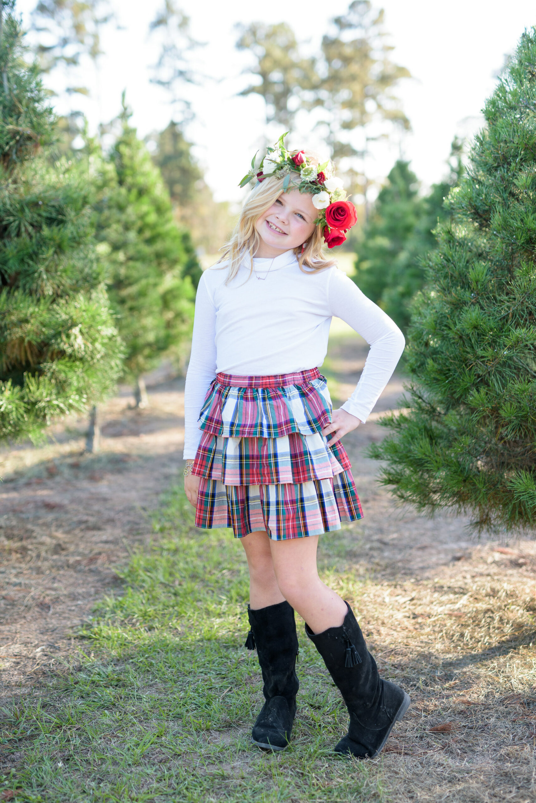 Christmas Tree Farm Photos by popular Houston lifestyle blog, Fancy Ashley: image of a girl standing in a row of pine trees and wearing a white long sleeve turtleneck shirt, plaid ruffle tier skirt, floral crown and black suede boots. 