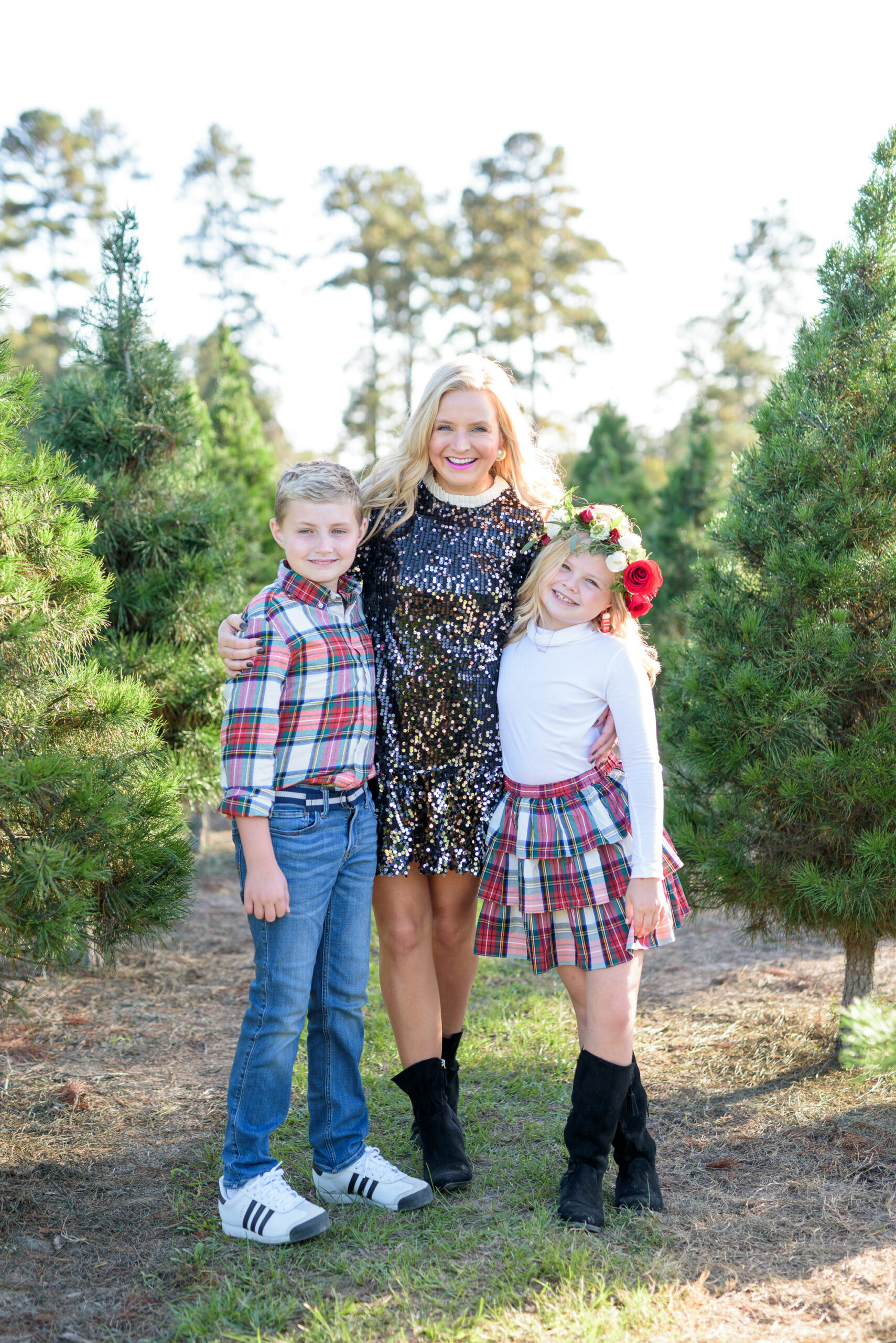 Christmas Tree Farm Photos by popular Houston lifestyle blog, Fancy Ashley: image of a mom and her two children standing together in a row of pine trees and wearing a black sequin dress, black suede ankle boots, plaid button up shirt, jeans, white long sleeve turtleneck shirt, plaid ruffle tier skirt, floral crown and black suede boots. 