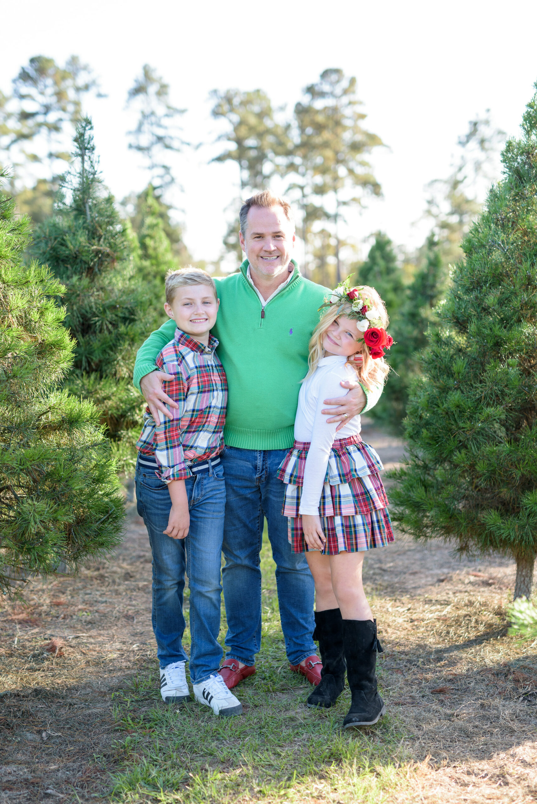 Christmas Tree Farm Photos by popular Houston lifestyle blog, Fancy Ashley: image of a dad and his two children standing together in a row of pine trees and wearing a green pullover sweater, red loafers, black suede boots, plaid button up shirt, jeans, white long sleeve turtleneck shirt, plaid ruffle tier skirt, floral crown and black suede boots. 