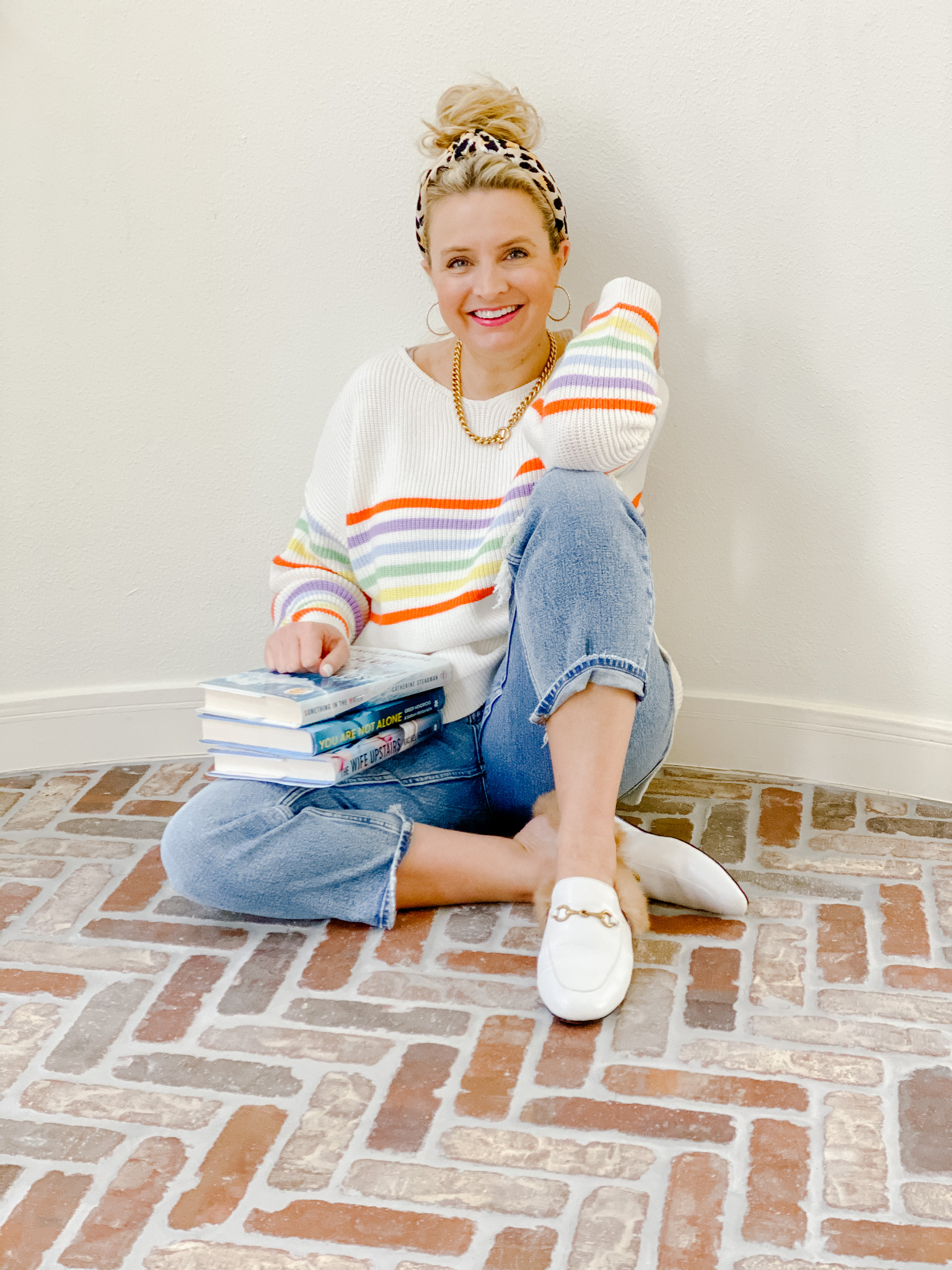 January Books by popular Houston lifestyle blog, Fancy Ashley: image of a woman wearing a multi strip sweater, light wash denim, white fur lined mules, leopard print knot headband, and gold hoop earrings, while sitting on a brick floor with a stack of books. 