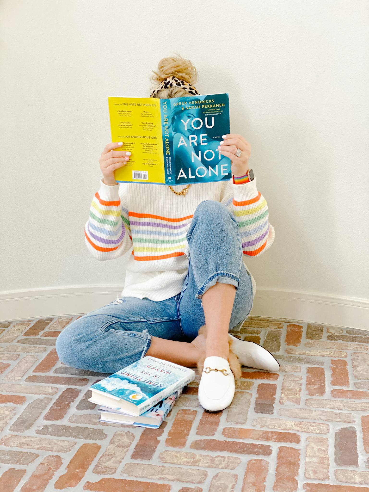 January Books by popular Houston lifestyle blog, Fancy Ashley: image of a woman reading the book You Are Not Alone while sitting on a brick floor and wearing a multi color stripe sweater, light wash jeans, fur lined white mules, and a leopard print knot headband. 