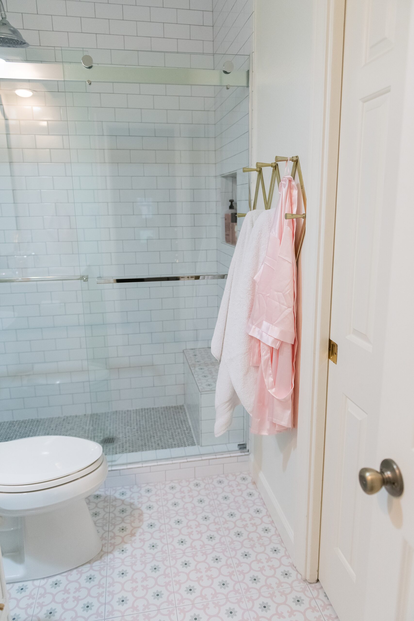 Girls Bathroom by popular Houston life and style blog, Fancy Ashley: image of a bathroom with a walk in shower that has white subway tile, pink and grey tiles, and grey penny tiles, pink and grey tile flooring a gold accordion wall hook with pink and white towels hanging from them. 