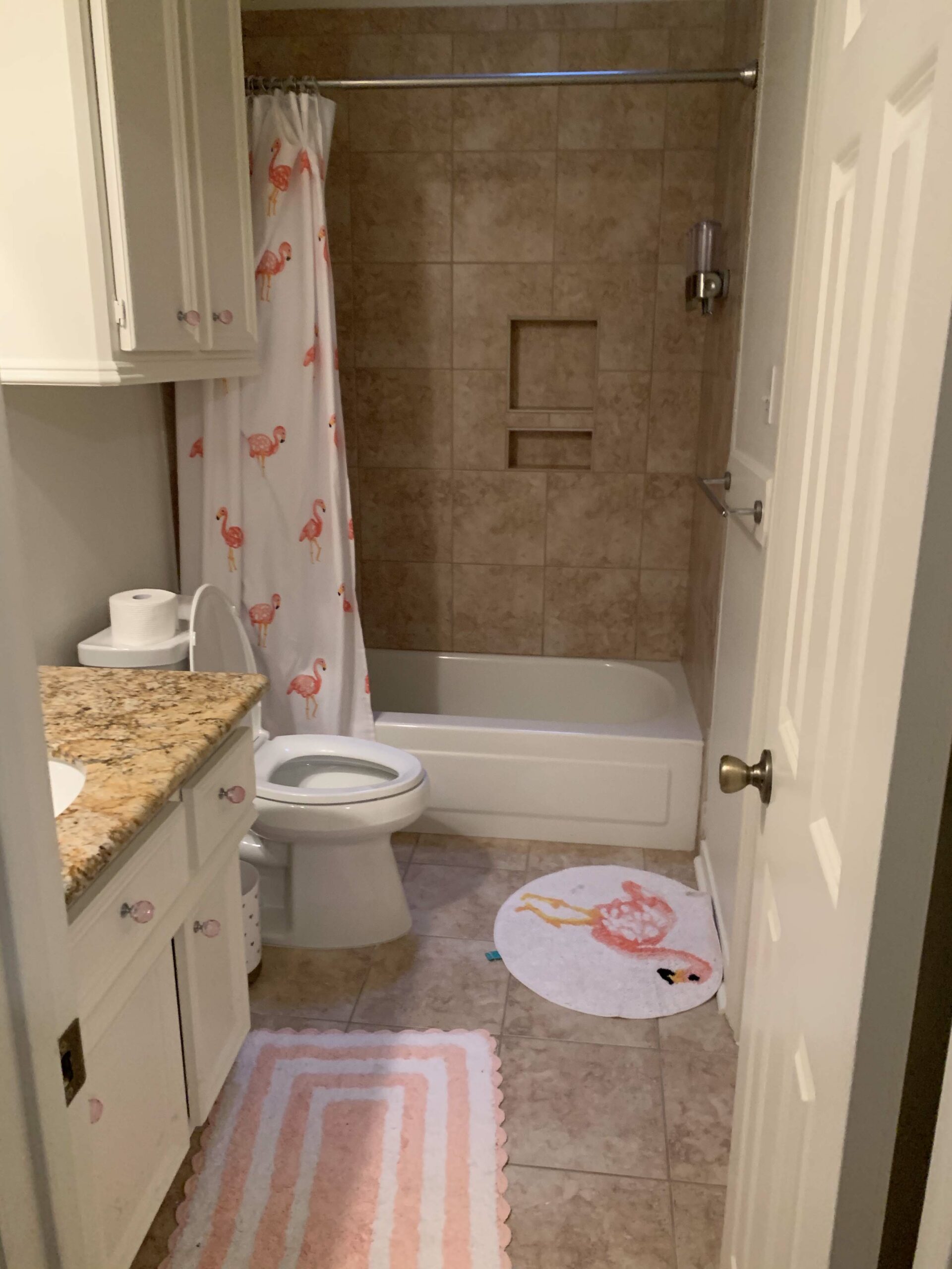 Girls Bathroom by popular Houston life and style blog, Fancy Ashley: image of a bathroom with brown tile, white cabinets, pink crystal drawer pulls, flamingo shower curtain, flamingo rug, and pink and white stripe scallop rug. 