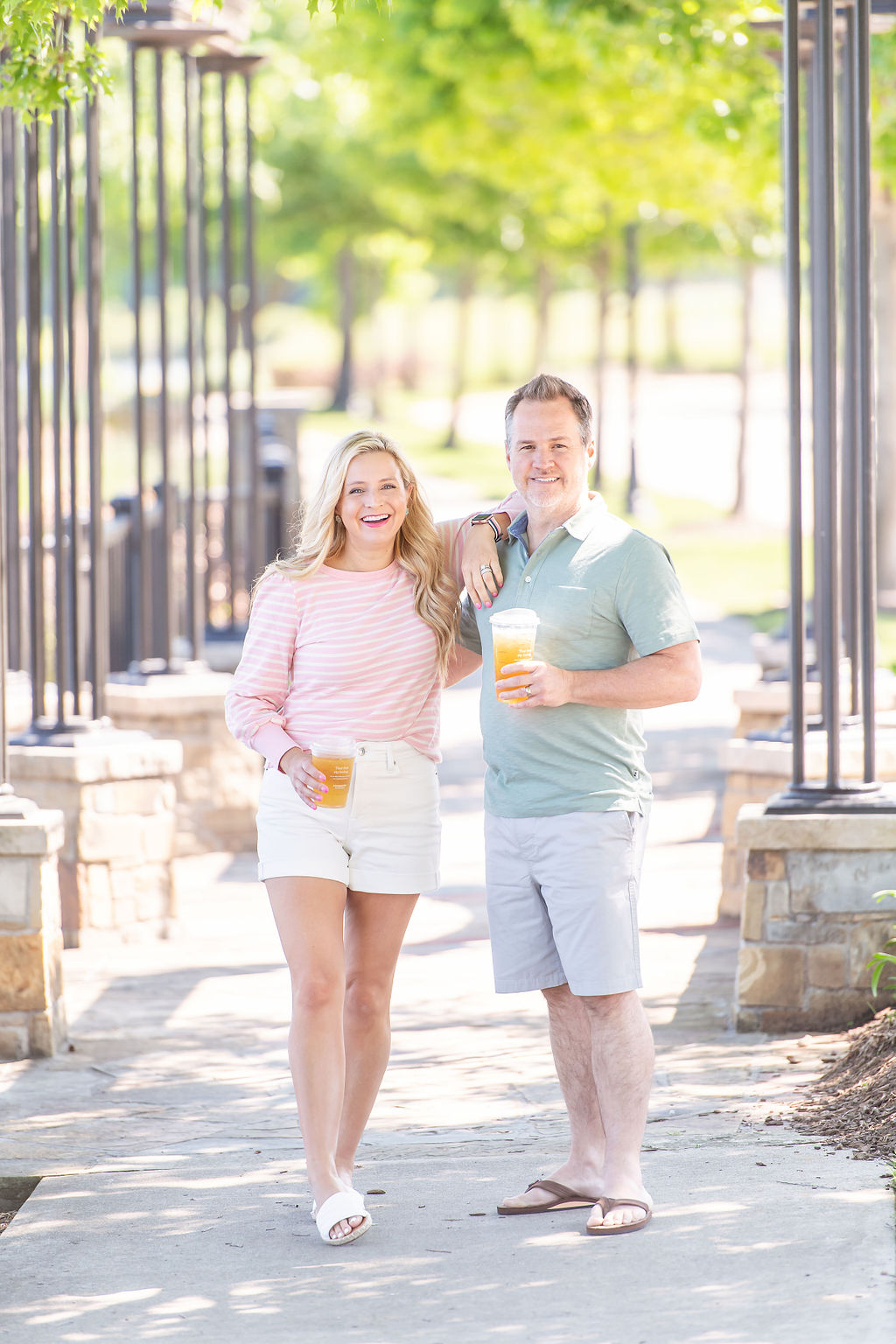 Walmart Summer Fashion by popular Houston fashion blog, The House of Fancy: image of a husband and wife standing together and wearing a pink and white stripe long sleeve top, white, shorts, white slide sandals, green polo, grey shorts and tan thong sandals. 