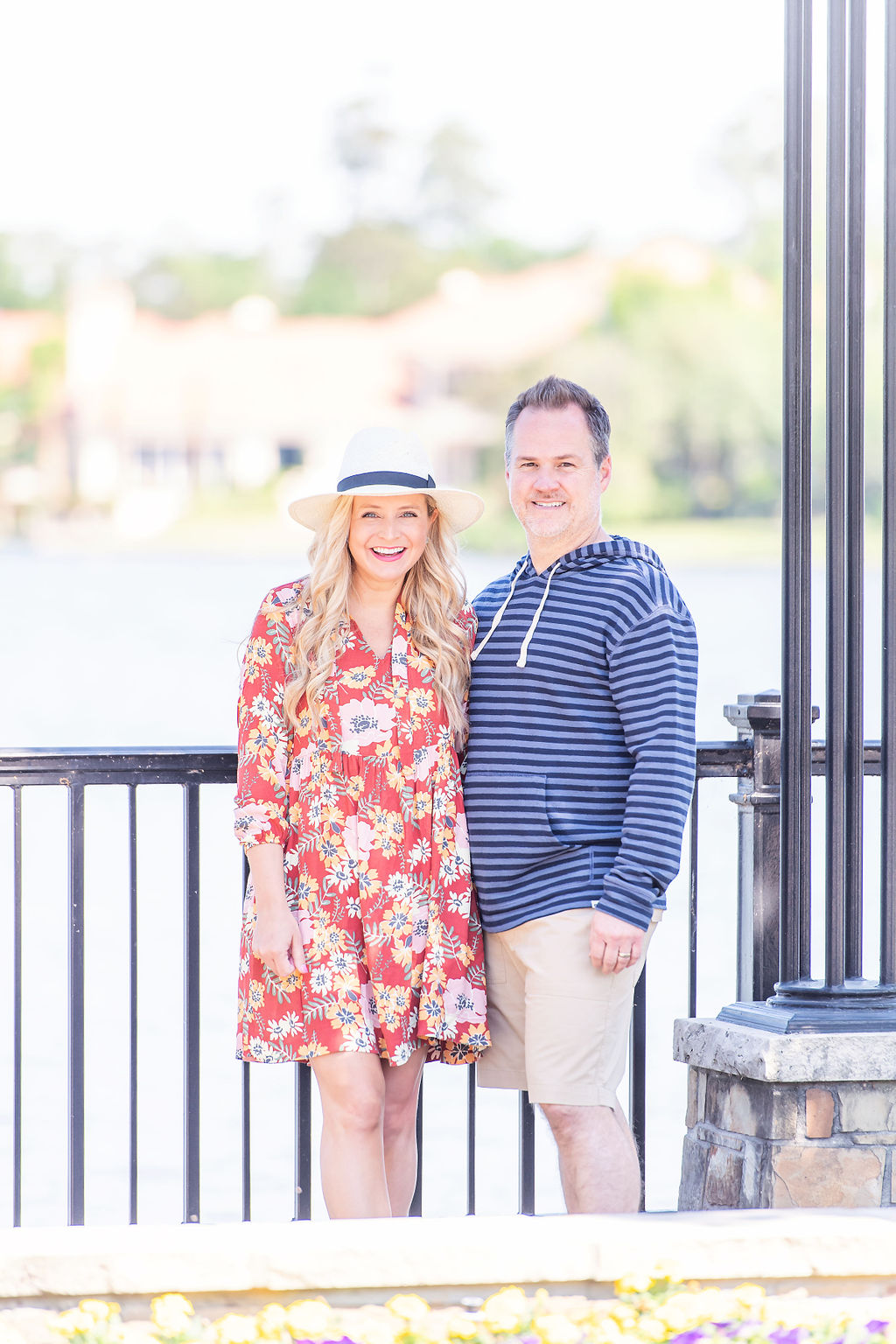 Inspire Others by popular Houston lifestyle blog, The House of Fancy: image of a husband and wife standing together outside by a black iron fence and wearing a red floral print dress, straw fedora hat, and blue stripe hoodie, tan shorts. 