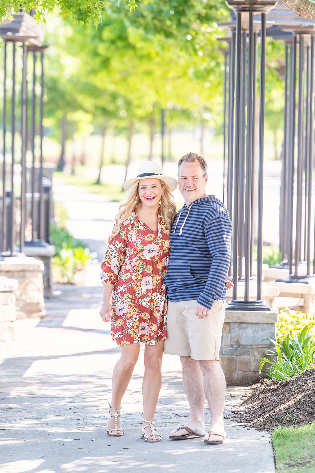 Walmart Summer Fashion by popular Houston fashion blog, The House of Fancy: image of a husband and wife standing together and wearing a red floral print dress, straw sun hat, studded gold strap sandals, brown thong sandals, blue stipe hoodie sweatshirt, and tan shorts. 