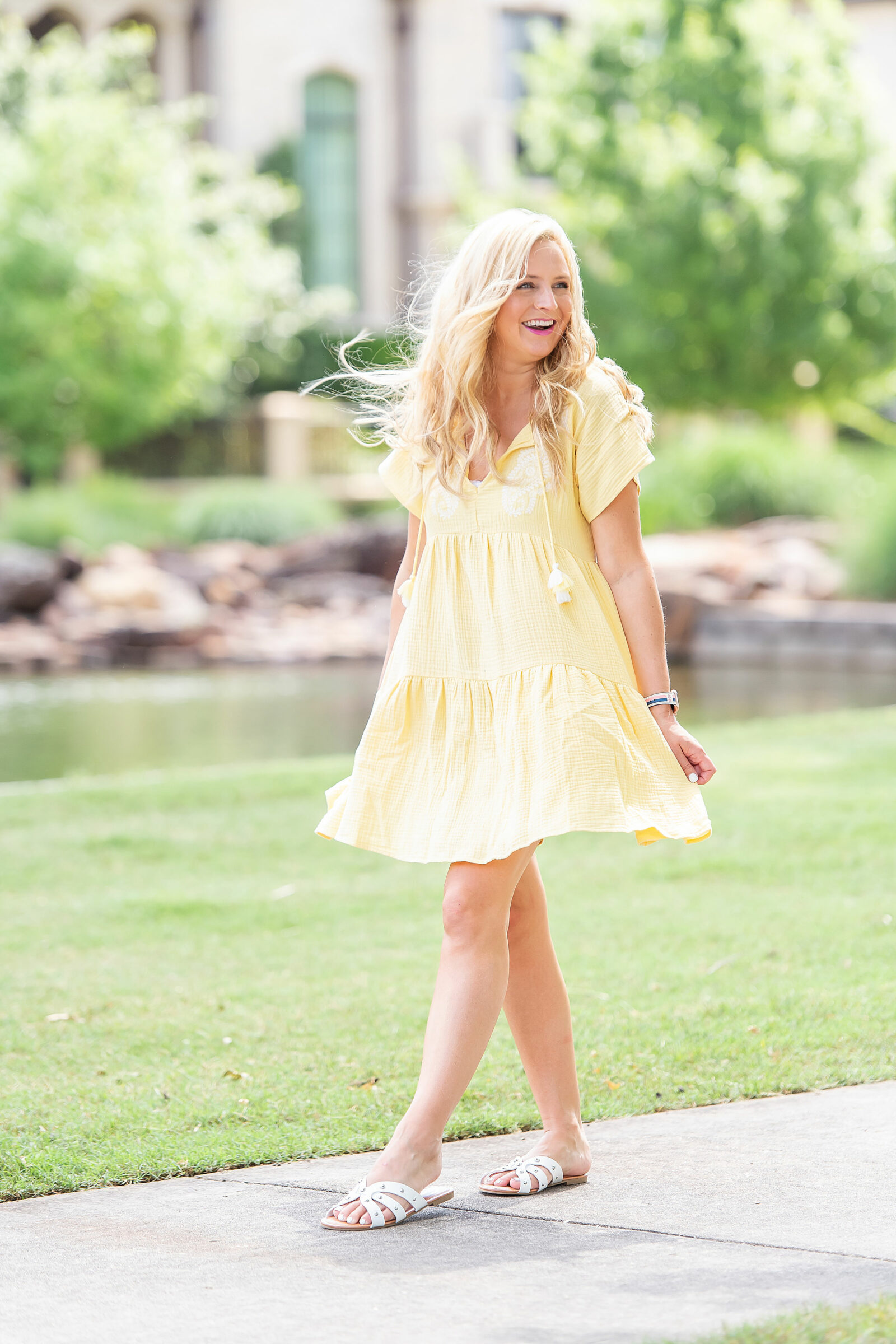 The Prettiest Embroidered Dresses for Summer |. Prettiest Embroidered Dresses for Summer | Embroidered Dresses by popular Houston fashion blog, The House of Fancy: image of a woman walking outside on a sidewalk and wearing a yellow embroidered dress with white strap slide sandals. 