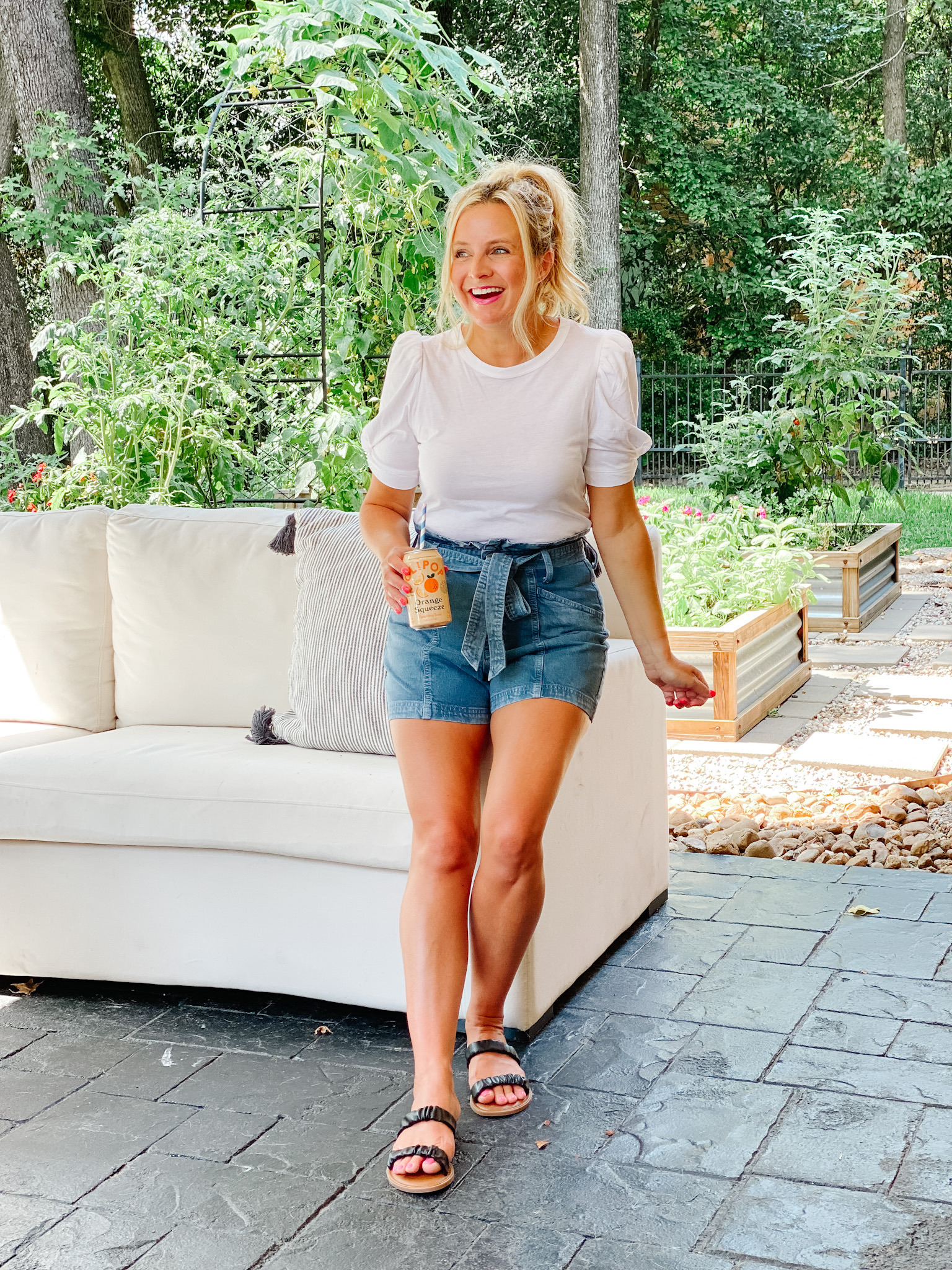 Summer Looks from Target by popular Houston fashion blog, The House of Fancy: image of a woman standing outside and leaning against a patio couch and holding a can of soda with a blue and white stripe straw in it and wearing a white puff sleeve t-shirt, paper bag denim shorts, and black strap sandals. 