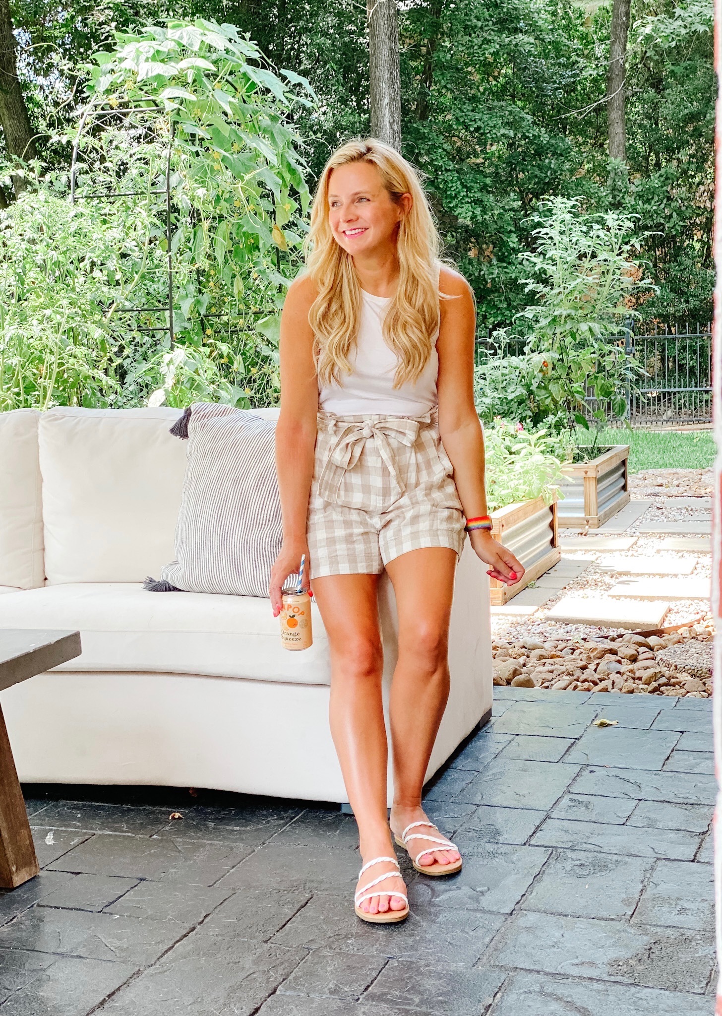 Summer Looks from Target by popular Houston fashion blog, The House of Fancy: image of a woman standing outside and leaning against a patio couch and holding a can of soda with a blue and white stripe straw in it and wearing a white tank top and tan and white buffalo plaid paper bag shorts with white strap sandals. 