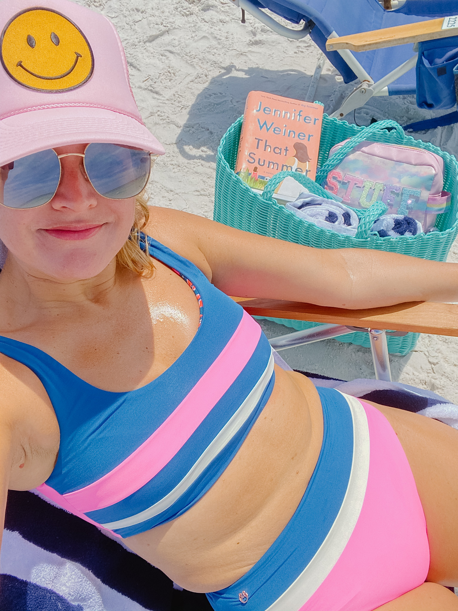 Best Beach Reads by popular Houston lifestyle blog, The House of Fancy: image of a woman sitting in a beach chair and wearing a blue, white and pink stripe two swimsuit, pink smiley face baseball hat, and sitting next to a turquoise tote bag filled with blue and white stripe towels and the book, That Summer. 