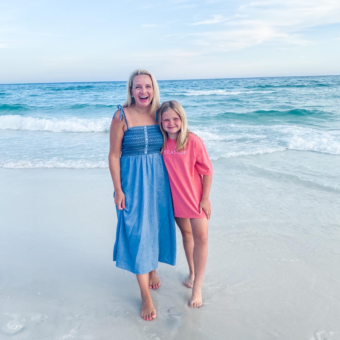 Seaside Florida by popular Houston travel blog, The House of Fancy: image of a mom and her young daughter standing together on a white sand beach next to the ocean and wearing a smocked tie strap denim dress and oversized pink Seaside t-shirt. 