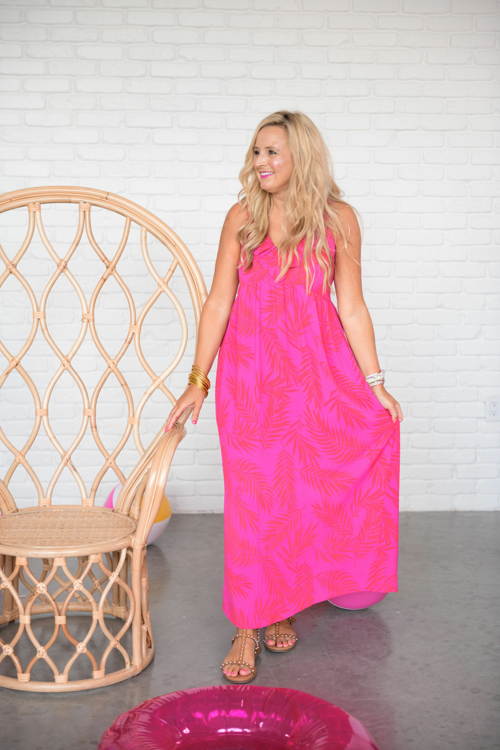 Summer Clothing from Gibson Look by popular Houston fashion blog, The House of Fancy: image of a woman standing next to a bamboo peacock chair and wearing a pink and red tropical print maxi dress with brown studded sandals.  